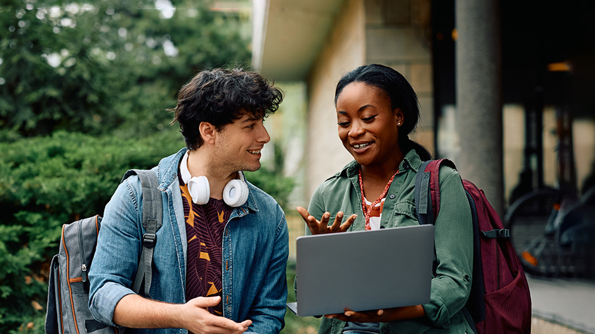 Two students walk around, smile whilst looking at a laptop screen