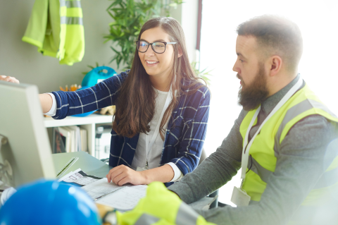 Industry_Marketing_Construction_Article_Text_Image_2_260324 Two construction workers, one a woman in a suit and the other a man in hi vis jacket, sat at a desk looking at a computer screen