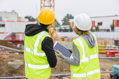 Industry_Marketing_Construction_Article_Text_Image_3_260324 Two construction workers, wearing hi vis jackets and helmets, stood outside looking at a construction site