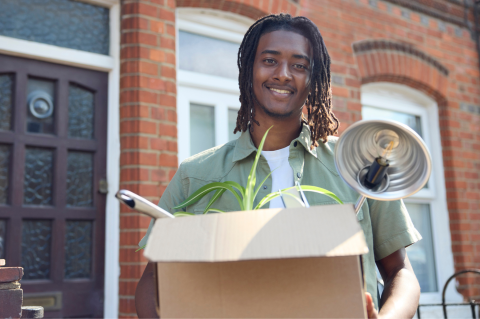 web-accommodation-image-box-2-2048-1361-080824.png A student stood outside the front door of a terraced house, holding a box containing a plant and lamp